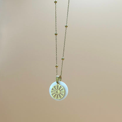 Goldfarbene Halskette auf lachsfarbenem Hintergrund. Die Kette hat kleinen Kugeln als Details und der Anhänger ist rund und zeigt eine Sonne in deren Zentrum ein Mond zu sehen ist. Der Anhänger liegt auf einem Perlmuttplättchen