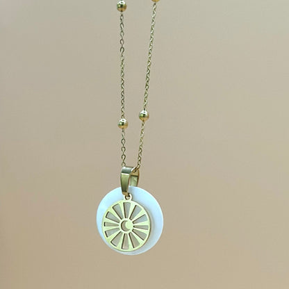 Goldfarbene Halskette auf lachsfarbenem Hintergrund. Die Kette hat kleinen Kugeln als Details und der Anhänger ist rund und zeigt eine Sonne in deren Zentrum ein Mond zu sehen ist. Der Anhänger liegt auf einem Perlmuttplättchen
