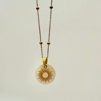 Goldfarbene Halskette auf weißem Hintergrund. Die Kette hat kleinen Kugeln als Details und der Anhänger ist eine Sonne, deren Strahlen kleine, aneinander gereihte Kugeln sind. Der Anhänger liegt auf einem Perlmuttplättchen.