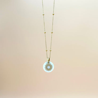 Goldfarbene Halskette auf lachsfarbenem Hintergrund. Die Kette hat kleinen Kugeln als Details und der Anhänger ist eine Sonne, deren Strahlen kleine, aneinander gereihte Kugeln sind. Der Anhänger liegt auf einem Perlmuttplättchen.