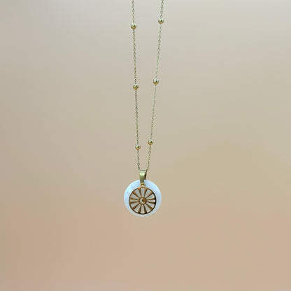 Goldfarbene Halskette auf lachsfarbenem Hintergrund. Die Kette hat kleinen Kugeln als Details und der Anhänger ist rund und zeigt eine Sonne in deren Zentrum ein Mond zu sehen ist. Der Anhänger liegt auf einem Perlmuttplättchen