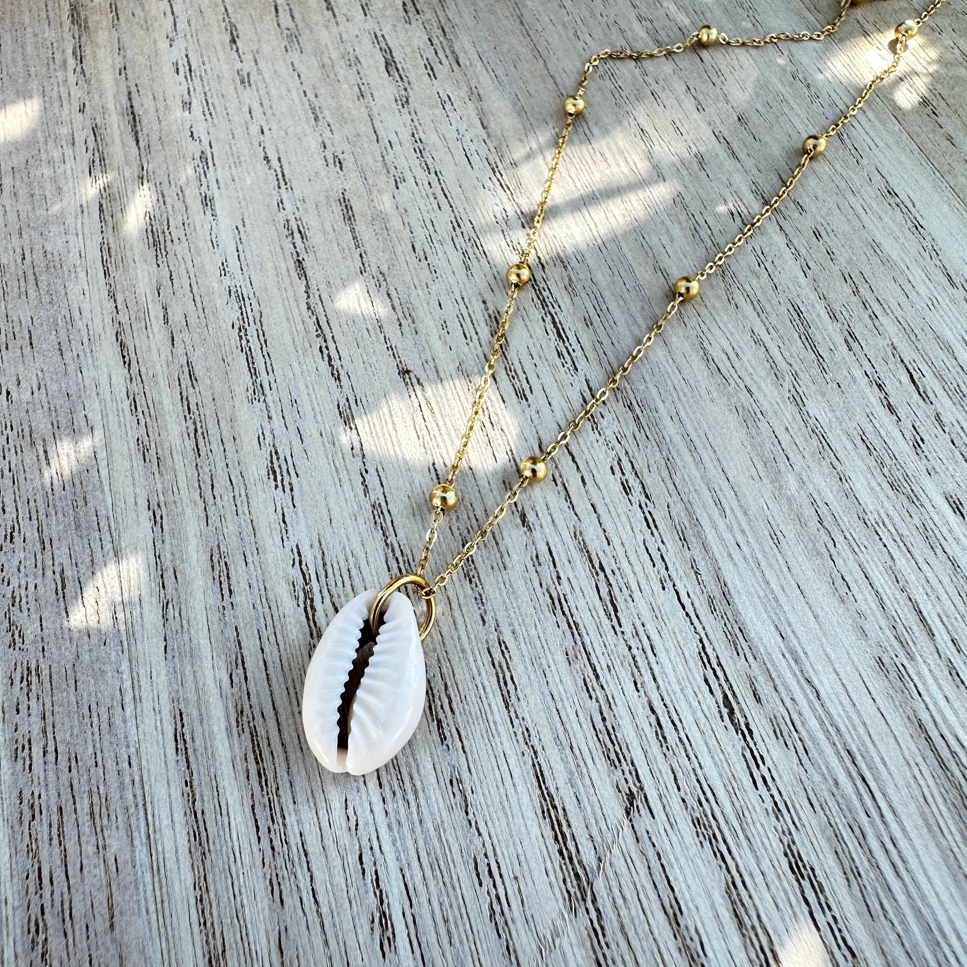 Weiße Kaurimuschel-Halskette mit goldener Edelstahlkette flachliegend auf Holztisch.